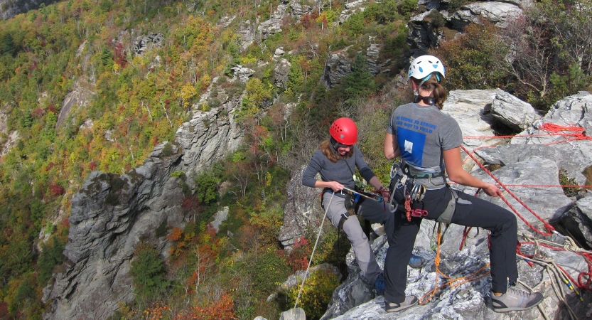 Two people wearing safety gear are secured by ropes near the edge of a cliff. 